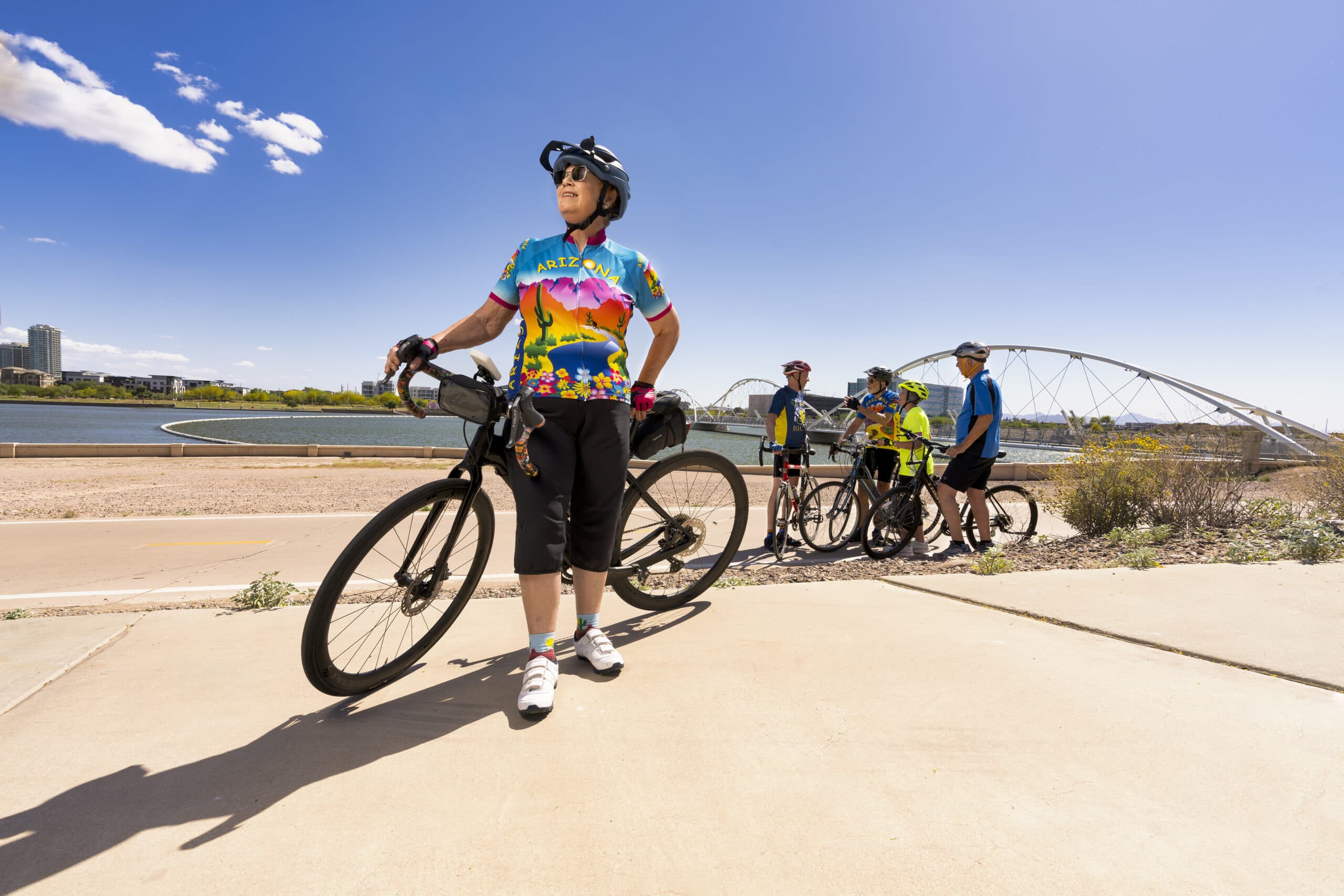 An older person stands with a bike in the foreground of the photo. A group of bikers stands in front of a picturesque lake in the background.