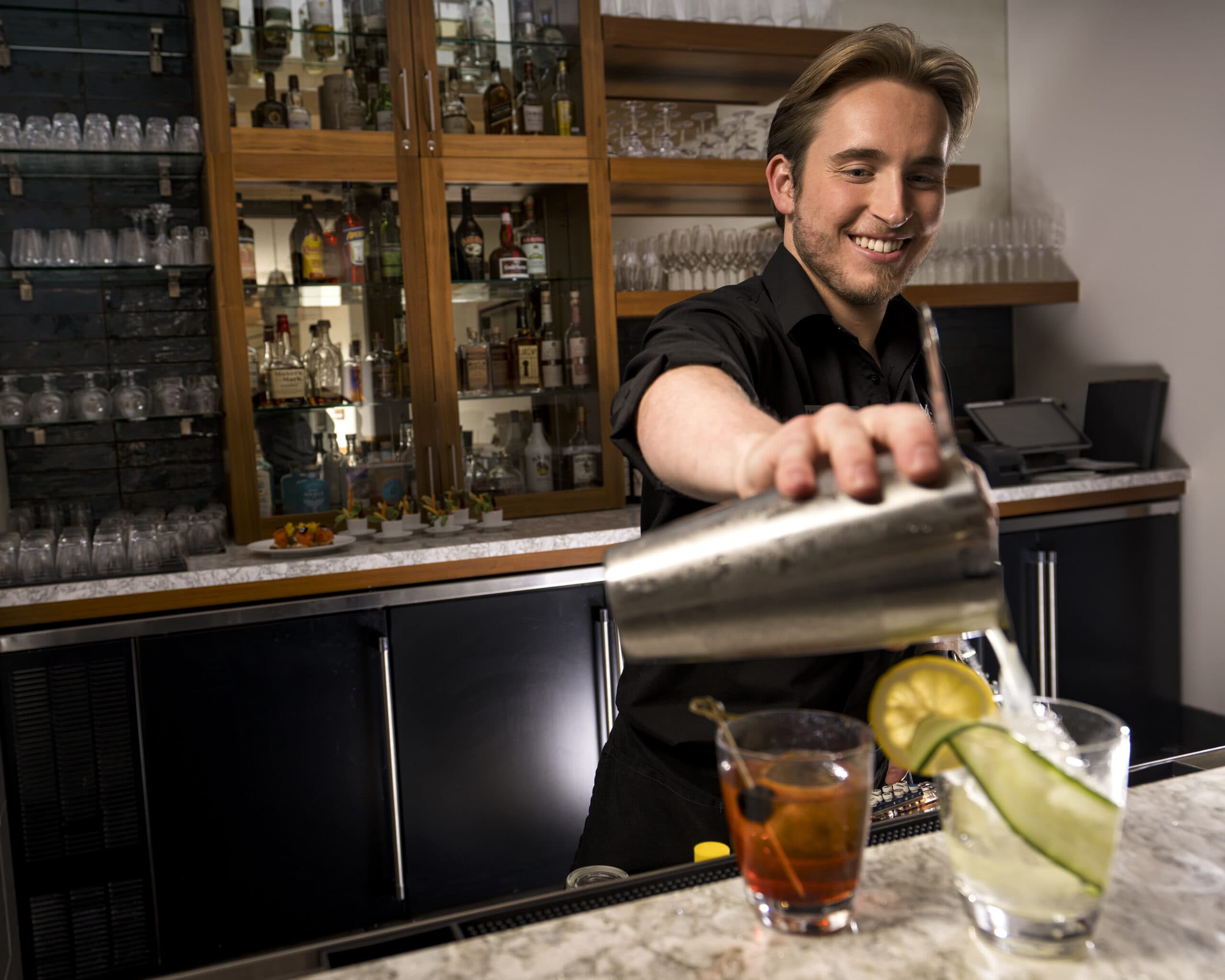 An expert mixologist pours a drink, garnished with lemon and cucumber.